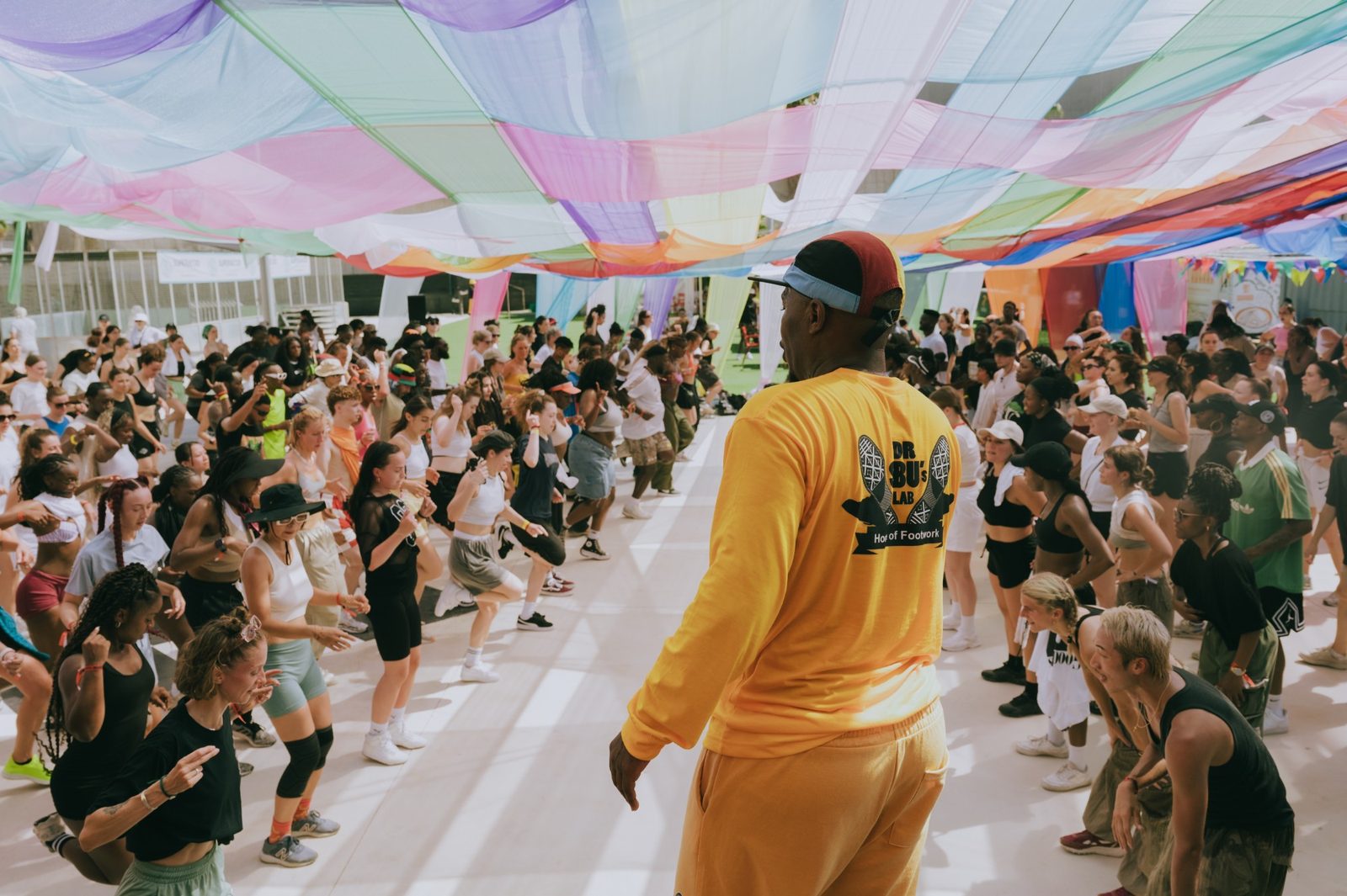 A teacher leading a large open-air class at OYOFE, hundreds of dancers following in rows under a canopy of coloured fabric